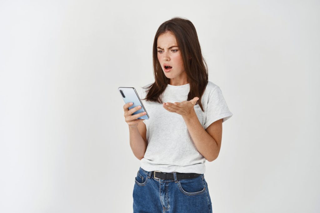 A young woman staring at her phone with her mouth open and one hand raised in frustration.