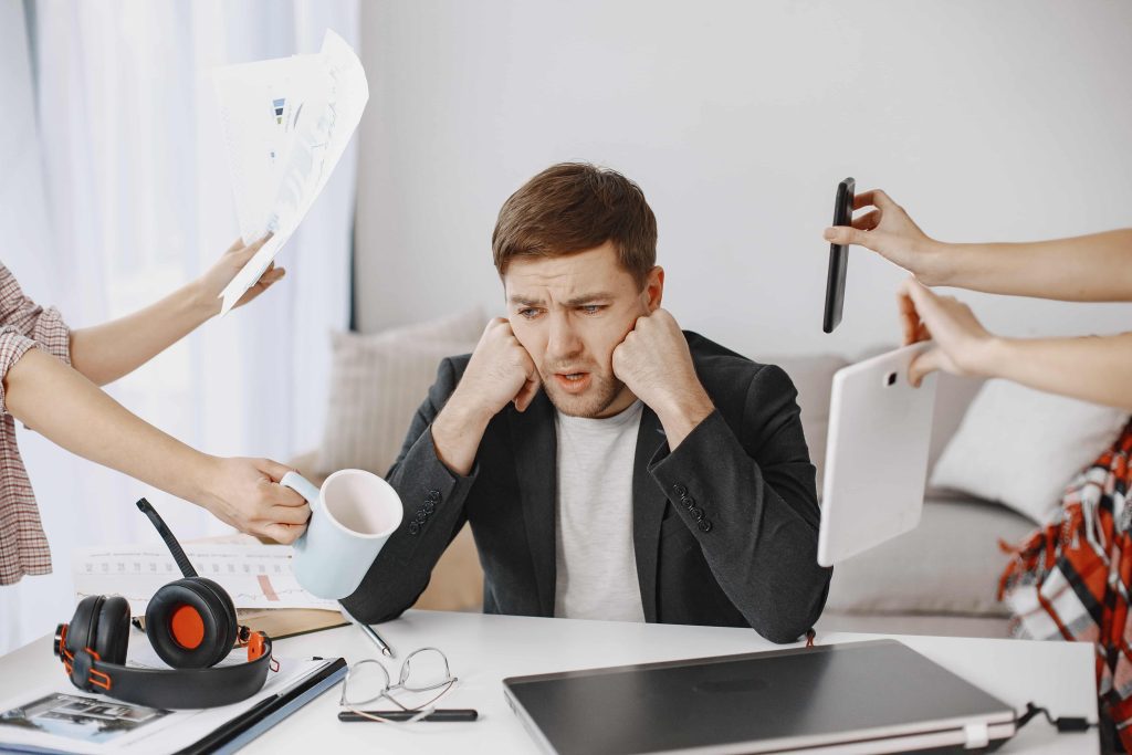 A stressed man at a desk, covering his ears while multiple hands offer him a mug, phone, tablet, and documents, symbolizing overwhelming multitasking and pressure at work.