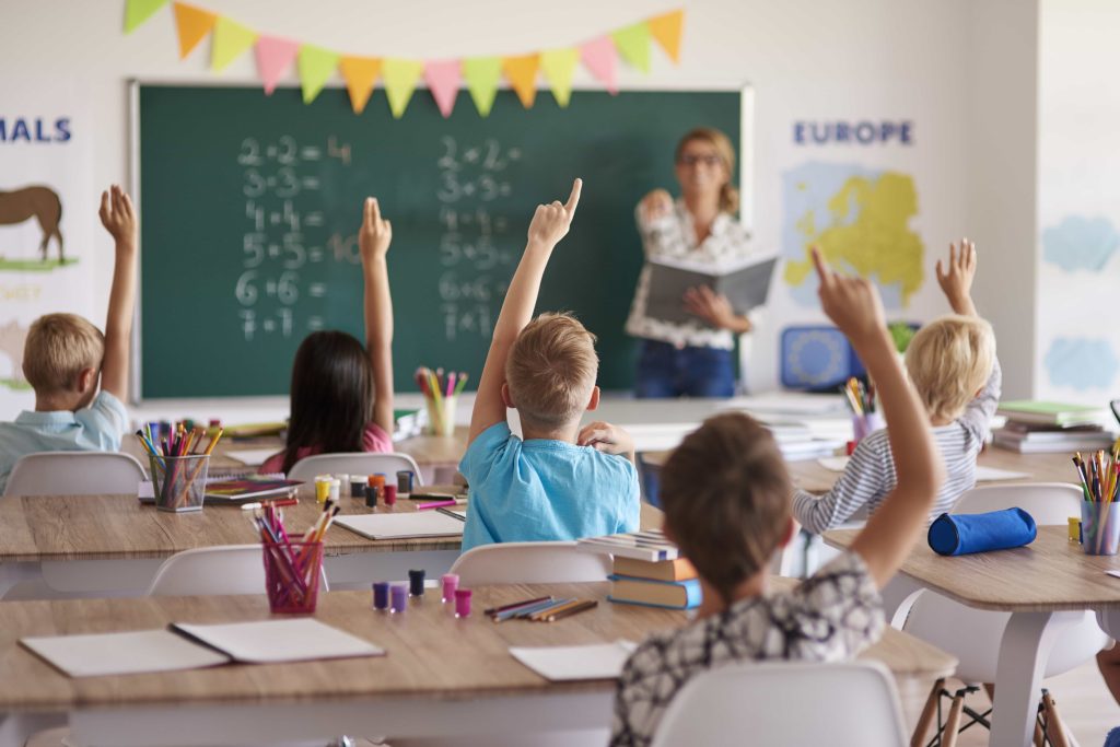 A classroom of young students with raised hands answering a teacher’s question in front of a chalkboard.