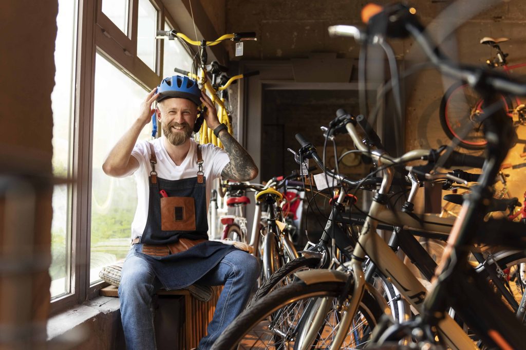 A smiling man in an apron and helmet inside a local bike shop surrounded by bicycles, showcasing a friendly and community-oriented small business.