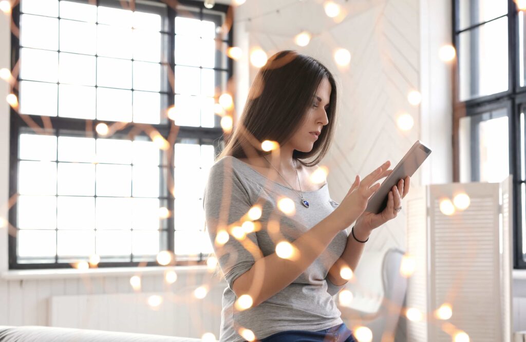 A woman in a gray off-shoulder shirt using a tablet indoors, with blurred festive string lights in the foreground.