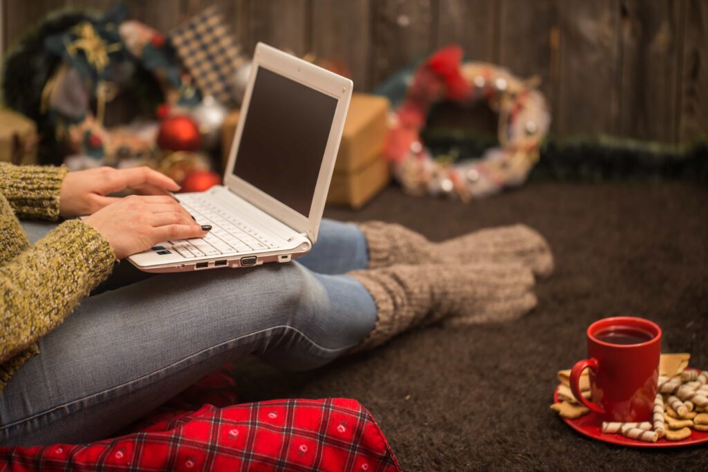 A person sitting on the floor with a laptop on their lap, wearing cozy socks, with a red mug of hot drink and festive Christmas decorations in the background.