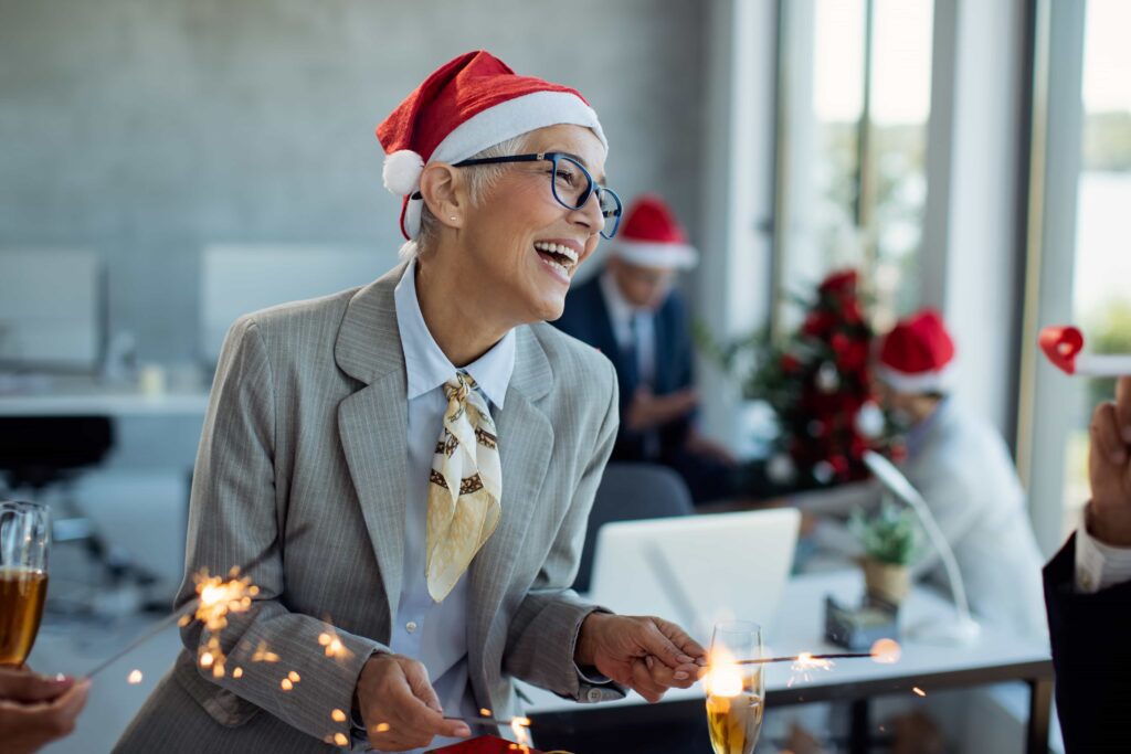 A cheerful mature woman wearing a Santa hat and business attire, holding sparklers and laughing at an office New Year's party, with colleagues and festive decorations in the background.