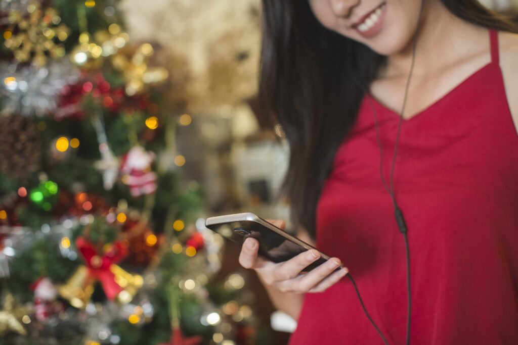 A woman in a red dress holding a smartphone and listening to music with earphones, standing near a brightly decorated Christmas tree with glowing lights.